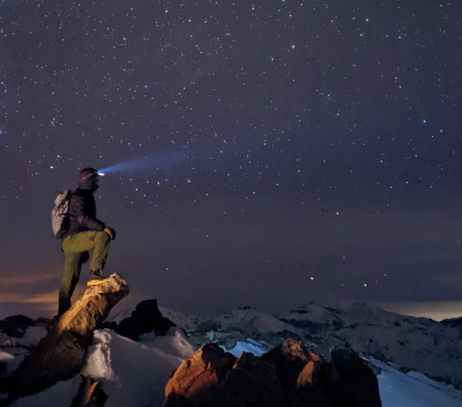 A hiker standing on a mountain beneath a star filled skys photographed using Google Pixels Night Sight mode